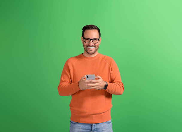孤立した緑の背景でスマートフォンを介してメッセージング眼鏡のハンサム笑顔ビジネスマン - 写真・画像