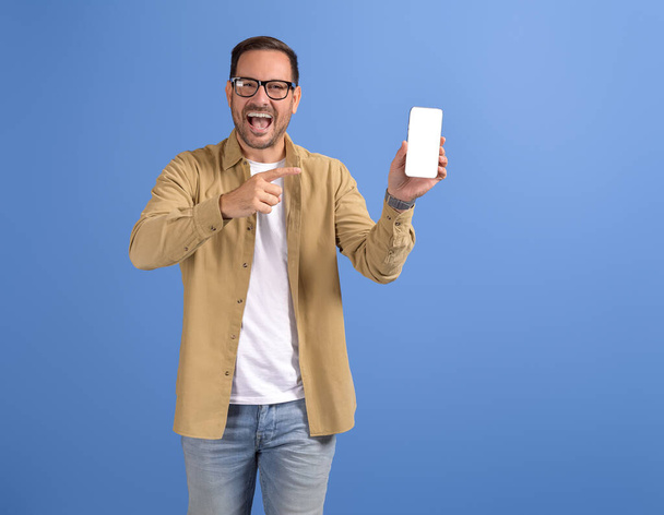 Πορτρέτο του χαρούμενου νέου άνδρα που δείχνει το smartphone και τη διαφήμιση που χρονολογείται app στο μπλε φόντο - Φωτογραφία, εικόνα