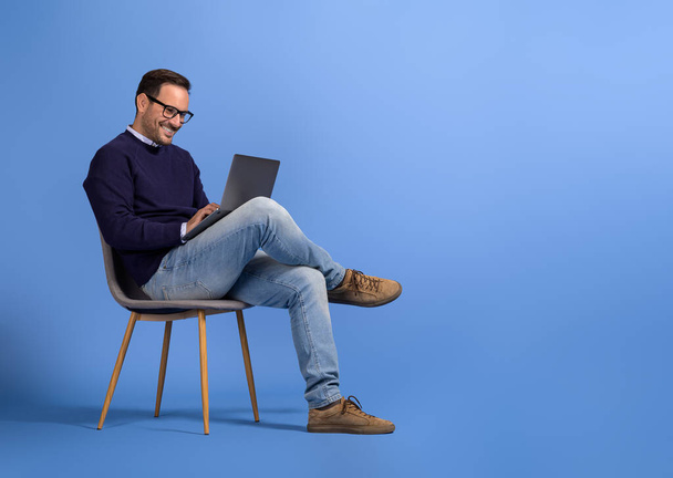 Pewny siebie młody biznesmen w okularach za pomocą laptopa podczas siedzenia na krześle na niebieskim tle - Zdjęcie, obraz