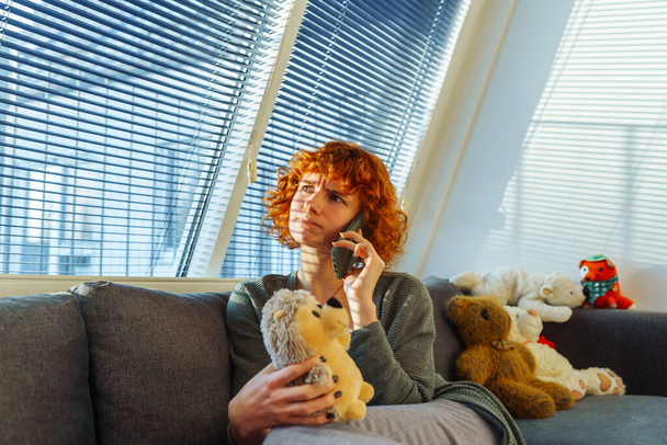 portrait red-haired curly teenage girl using phone while sitting on comfortable sofa at home, near large window with blinds. hugs childrens toys, nostalgic emotion. shadows from blinds fall on face - Photo, Image