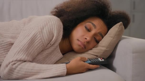 Kanepede tek başına uzanan Afrikalı Amerikalı yorgun kadın uzaktan kumandalı kanalları tutuyor sıkıcı yalnız hafta sonu televizyon seyrediyor sıkılmış tembel, üzgün, depresyondaki, ilgisiz bir kadın. - Video, Çekim