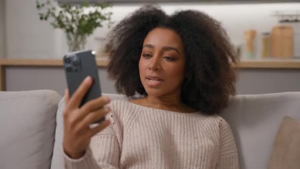 Afro-Amerikalı kadın, kaygısız, etnik bayan ev sahibi kanepe üzerinde gülümsüyor video konuşması, akıllı telefon, ev telefonuyla uzaktan iletişim kuruyor. - Video, Çekim