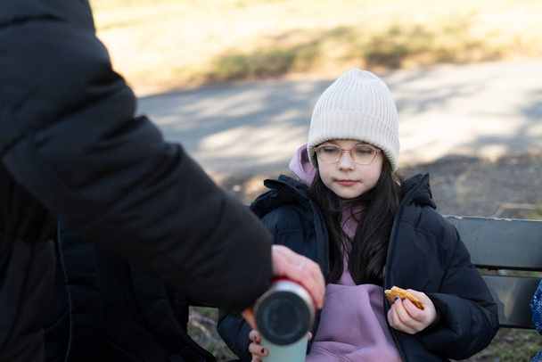 Маленькая девочка сидит на скамейке в парке со своим отцом и ест печенье. - Фото, изображение