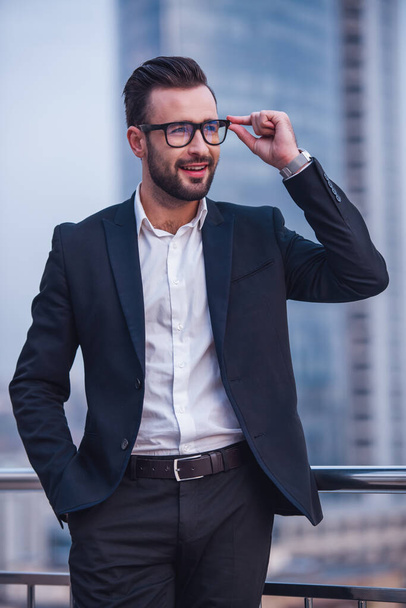 Pohledný podnikatel v obleku a brýle se dívá pryč a usměvavá stoje venku - Fotografie, Obrázek
