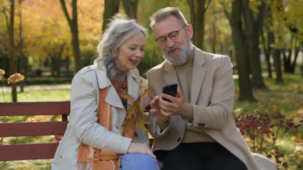 Caucasian retired couple family in love mature woman man sit on bench autumn park using mobile phone gadget together smiling wife husband spouses reading news old age and smartphone in city outdoors - Footage, Video