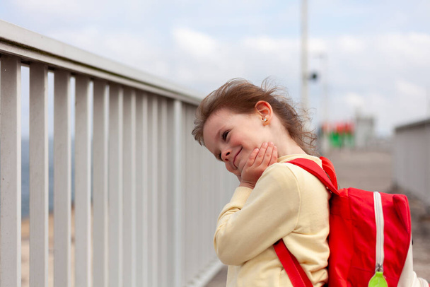 Ένα νεαρό κορίτσι με ένα κόκκινο σακίδιο στο δρόμο. Το κορίτσι είναι έκπληκτο.. - Φωτογραφία, εικόνα