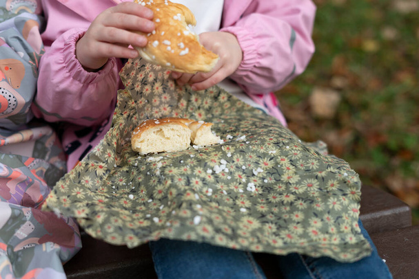 Petite fille assise sur un banc dans le parc et mangeant un pain fraîchement cuit - Photo, image