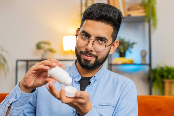 Indian young bearded man taking medicine pill with water sitting on cozy couch at home. Hispanic guy takes medication prescribed by his physician. Medicine, health care, pharmacy and people concept - Photo, Image