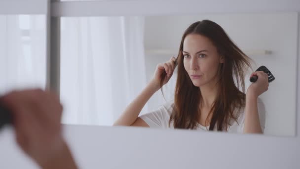 Nuori nainen käyttää curling pihdit muotoilla hiuksiaan katsoessaan peiliin valoisassa huoneessa, mikä heijastaa kauneutta ja grooming rutiini. - Materiaali, video