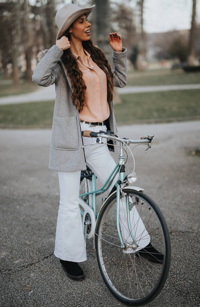 Elegante profesional femenina en un traje elegante posando con su bicicleta vintage en un entorno de parque de la ciudad. - Foto, Imagen