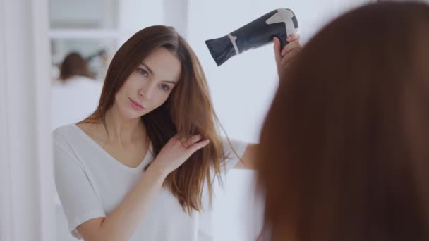 カジュアルなTシャツの女性は明るく照らされた部屋の鏡の前で吹くドライヤーを使用して髪をスタイルします. - 映像、動画