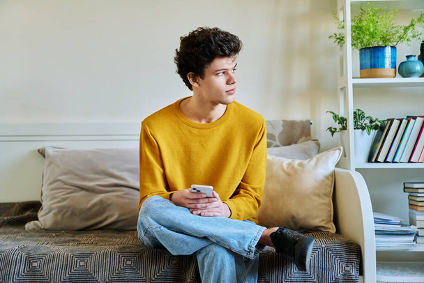 Young guy holding smartphone in hands sitting on couch at home. Handsome university college student 19-20 years old in yellow with curly hair. Technologies, mobile apps, study communication leisure - Foto, Imagen