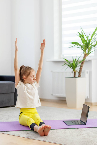 Yoga yaparken kollarını kaldıran yoga minderine odaklanmış ergen kız rahat ev ortamında dizüstü bilgisayar dersini takip ediyor.. - Fotoğraf, Görsel