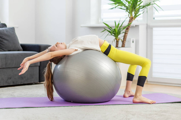 Dziewczyna używa piłki fitness do ćwiczeń rozciągających plecy, leżąc z tyłu z ramionami wyciągniętymi na macie do jogi w domu. - Zdjęcie, obraz