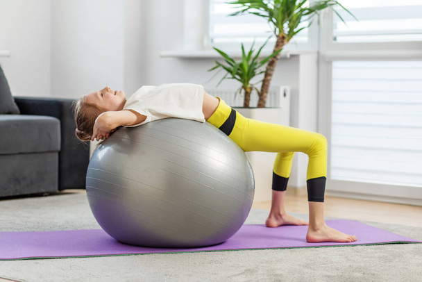Jong meisje voert backbend op fitness bal, ontspannen terwijl het uitrekken van haar rugspieren op paarse yoga mat thuis. - Foto, afbeelding