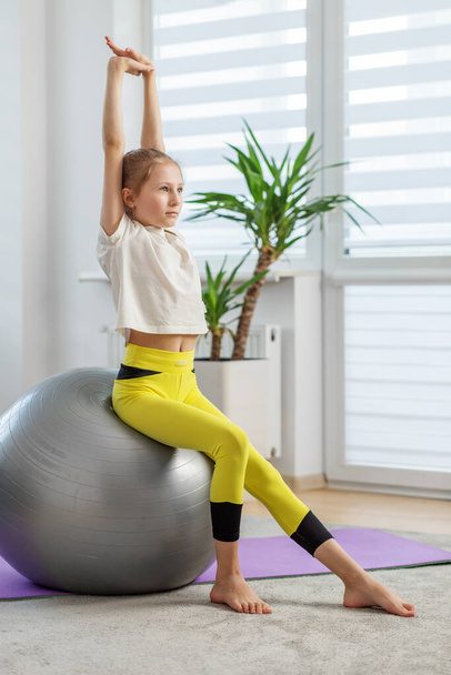 Nastoletnia dziewczyna siedząca na piłce fitness rozciąga ramiona, pracując nad swoją postawą i równowagą na fioletowej macie do jogi w dobrze oświetlonym pomieszczeniu. - Zdjęcie, obraz