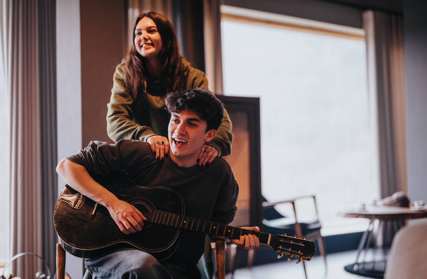 Radostný pár doma s mužem hrajícím na kytaru a ženou stojící za ním, usmívající se a bavící se. - Fotografie, Obrázek