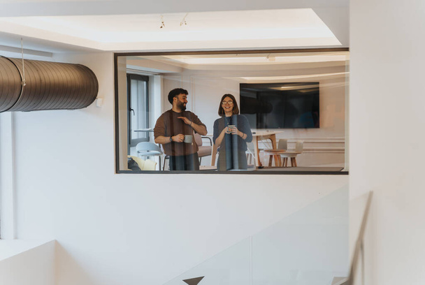 Een man en vrouw genieten van een luchtig moment met koffie in een stijlvolle, eigentijdse kantooromgeving, een weerspiegeling van kameraadschap op de werkplek. - Foto, afbeelding