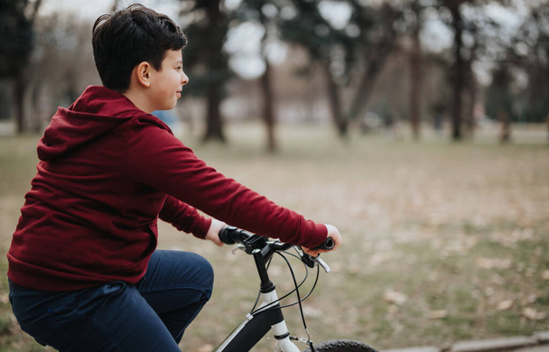 Ο ανέμελος νεαρός ποδηλατεί σε μονοπάτι μέσα σε αστικό πάρκο, περιτριγυρισμένος από πράσινο και απολαμβάνοντας τον καθαρό αέρα. - Φωτογραφία, εικόνα