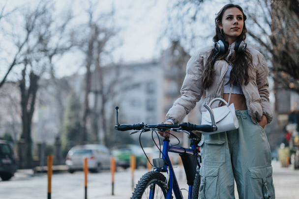 Pewna siebie młoda kobieta z rowerem zatrzymująca się na ulicy miejskiej, ubrana w luźny strój i słuchawki, ucieleśniająca miejski styl życia i rozrywkę. - Zdjęcie, obraz