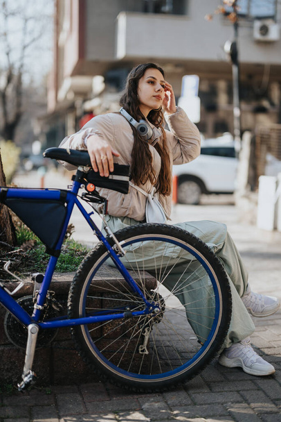 Młoda kobieta ze słuchawkami na szyi, korzystająca ze smartfona siedzącego przy niebieskim rowerze na miejskiej ulicy. - Zdjęcie, obraz