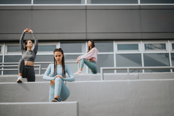 Три молоді жінки в спортивному одязі розтягуються і готуються до тренування на бетонних сходах у місті. - Фото, зображення