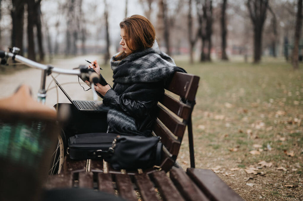Elegáns és magabiztos fiatal nő elegáns öltözékben, aki a munkájába van foglalva egy laptopon, miközben egy padon ül a parkban.. - Fotó, kép