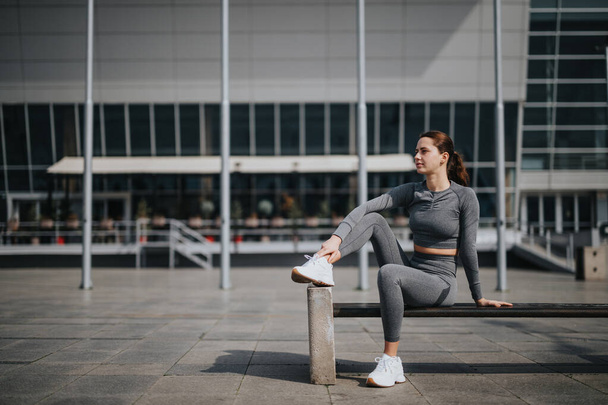 Zdolna młoda kobieta w odzieży sportowej spoczywa na ławce z nowoczesnym budynkiem w tle, ucieleśniającym zdrowy styl życia. - Zdjęcie, obraz