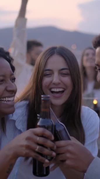 Vertical. Group of young diverse people dancing excited and toasting with beers at rooftop barbecue party. Laughing friends jumping and having fun drinking at event in sunset summer. Slow motion - Footage, Video
