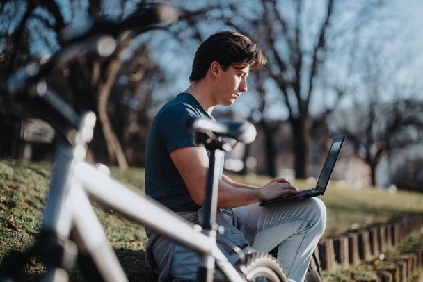 Ένας εστιασμένος άντρας κάθεται σε ένα παγκάκι με ένα ποδήλατο δίπλα του, ασχολούμενος με τη δουλειά στο λάπτοπ του σε ένα πάρκο.. - Φωτογραφία, εικόνα