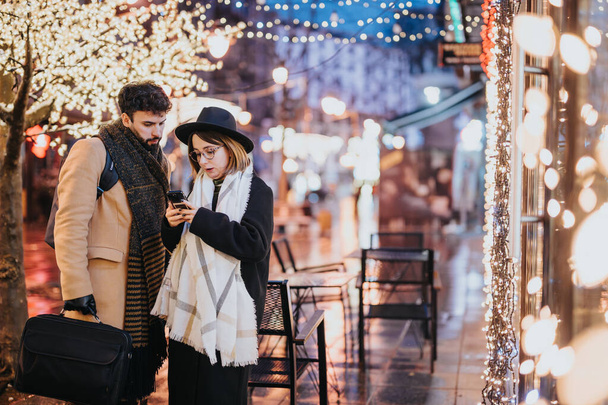 jong paar met behulp van smart phone op feestelijke stad straat versierd met kerstverlichting. - Foto, afbeelding