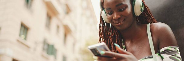 Gorgeous woman with African braids wearing top sits with headphones outside on the street and uses mobile phone, Panorama. Stylish girl listening to music on smartphone - Foto, Imagen