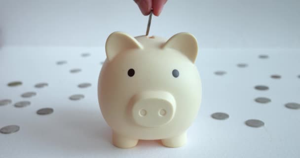 hand carefully places coin into piggy bank white table, amidst other coins, savings. symbol financial prudence, underlines importance accumulating savings for future security savings financial health - Séquence, vidéo