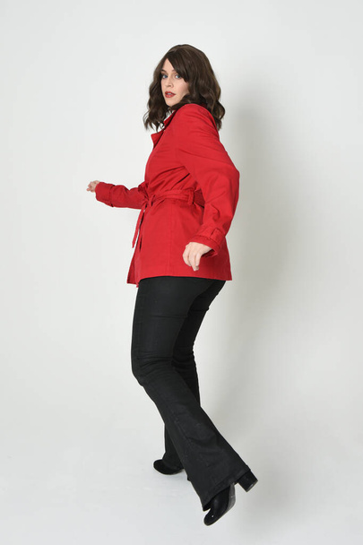 πλήρους μήκους πορτρέτο του όμορφη μελαχρινή γυναίκα μοντέλο, φορώντας κόκκινο σακάκι καμπαρντίνα δερμάτινο παντελόνι. Στέκεται ποζάρουν στο παρασκήνιο, με τα πόδια από την κάμερα. απομονωμένη σιλουέτα σε λευκό φόντο στούντιο. - Φωτογραφία, εικόνα