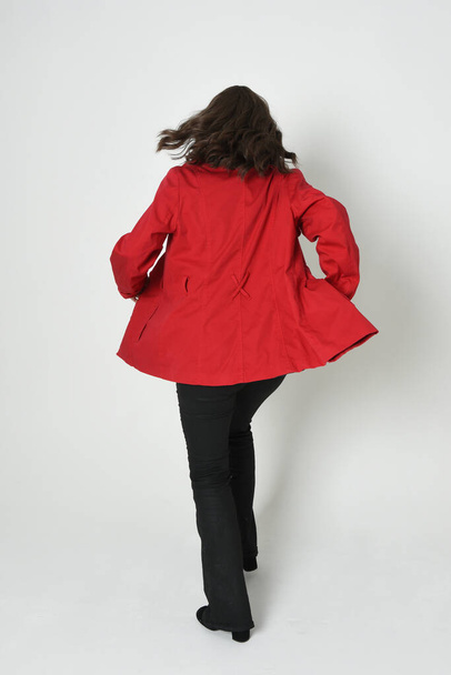 美しいブルネットの女性モデルの完全な長さの肖像画は,赤いトレンチコートジャケットの革のズボンを着用しています. バックビューに立って カメラを離れて。 ホワイトスタジオの背景にある孤立したシルエット. - 写真・画像