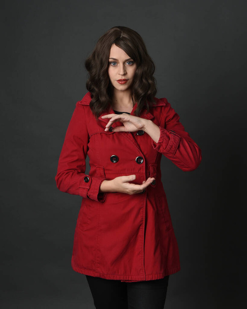 zbliżenie portret pięknej brunetki model kobiety, noszenie czerwony płaszcz okop z gestural ręce pozing.isolated na ciemnym tle studio z cieniami.  - Zdjęcie, obraz