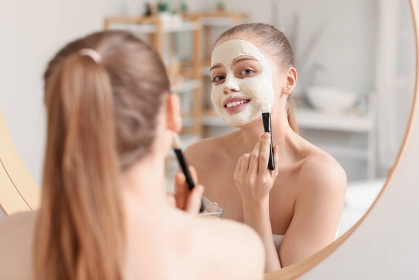 Jeune femme appliquant masque de curcuma avec brosse près du miroir à la maison - Photo, image