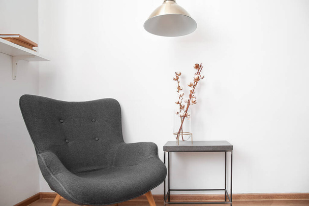 Vase aus Baumwollzweigen auf schwarzem Tisch mit Lampe und Sessel neben weißer Wand - Foto, Bild