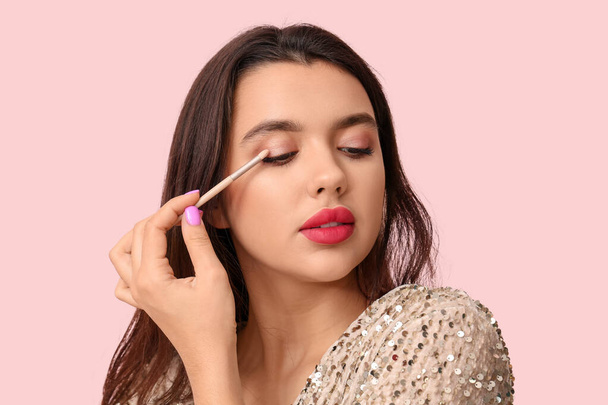 Beautiful young woman with brush applying eyeshadows on pink background - Photo, image