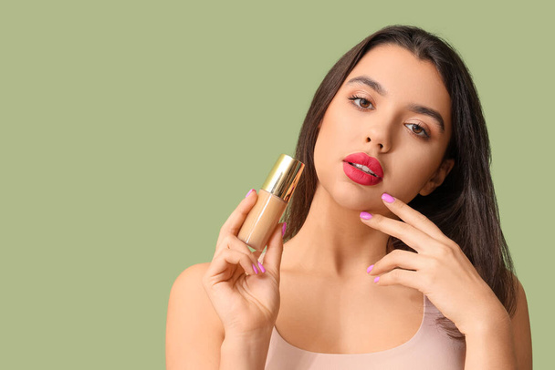 Beautiful young woman with bottle of makeup foundation on green background - Foto, imagen
