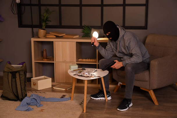 Мужчина-вор с пистолетом и украденными ценностями на столе в комнате ночью - Фото, изображение