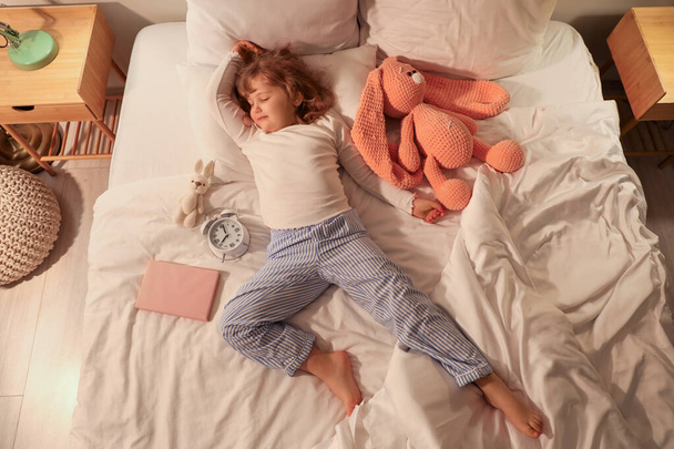 Rozkošná holčička s plyšovými hračkami, knihou a budíkem spící v posteli - Fotografie, Obrázek