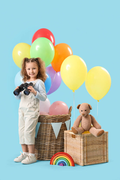 Nettes kleines Mädchen mit Fernglas, Körben mit Luftballons und Spielzeug auf blauem Hintergrund - Foto, Bild