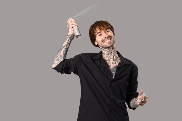 Jeune coiffeur tatoué avec spray capillaire sur fond gris - Photo, image