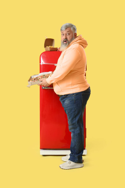 Homem maduro com sobrepeso chocado com pizza perto da geladeira em fundo amarelo. Conceito de excessos - Foto, Imagem
