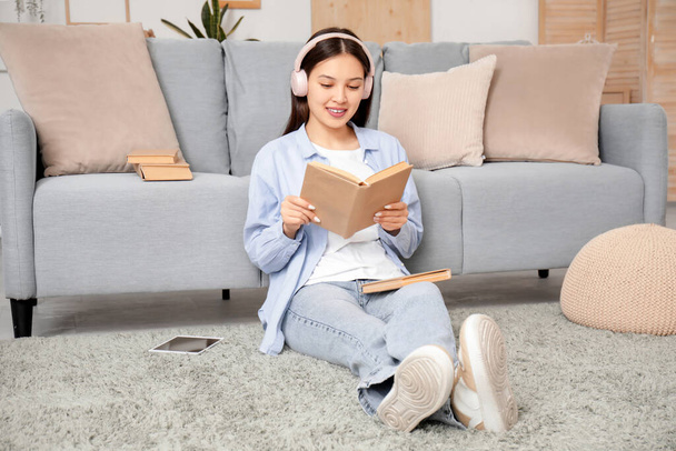 Kaunis nuori aasialainen nainen kuulokkeissa kuuntelemassa äänikirjaa kirjan kanssa olohuoneessa - Valokuva, kuva