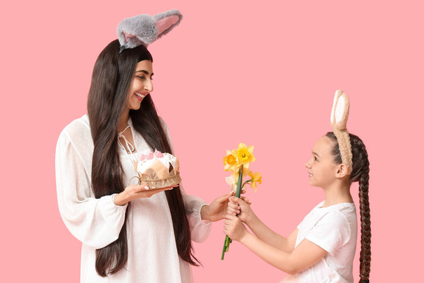 Happy smiling young woman with her daughter in bunny ears holding Easter cake and flowers on pink background - Photo, Image
