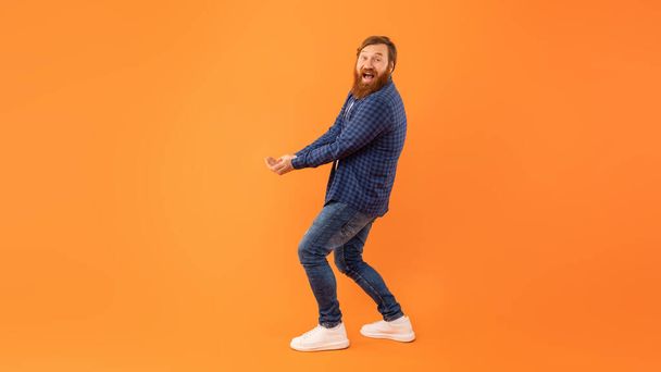 Prise de vue complète d'un homme aux cheveux roux positifs avec barbe tenant les mains ensemble avec les paumes levées, portant ou essayant d'attraper quelque chose d'invisible, debout au-dessus d'un mur de studio orange, panorama, espace de copie - Photo, image