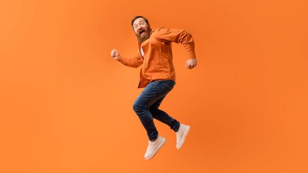 Nadšený zrzavý vousatý muž poskakující ve vzduchu a křičící vzrušením, bavící se na oranžovém pozadí studia, dívající se na kameru, vyjadřující šílené chladné vibrace. Panorama, volné místo - Fotografie, Obrázek