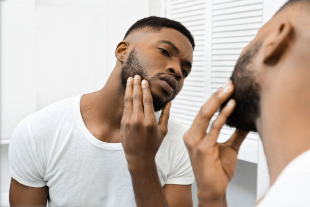 Africký Američan se dotýká své tváře, zatímco pečlivě zkoumá svou kůži, pravděpodobně hledá vady nebo nedokonalosti - Fotografie, Obrázek